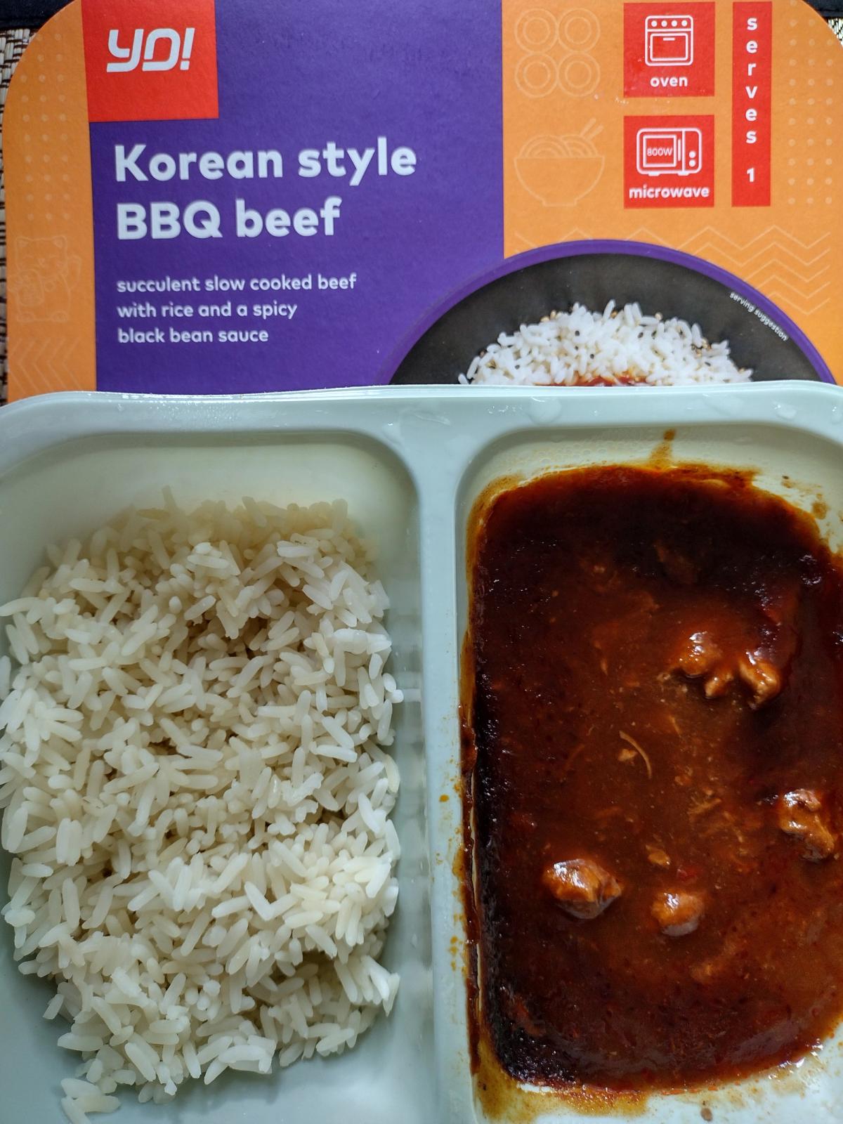 A two-compartment white plastic tray holding a meal. The left side contains cooked white rice, and the right side contains chunks of beef in a dark, glossy, spicy sauce. The purple YO! Korean style BBQ beef cardboard sleeve is in the background.