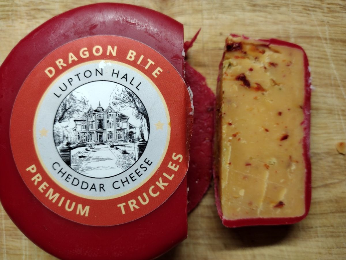 A wheel of Lupton Hall Dragon Bite spicy cheddar cheese in red wax, with a slice cut out on a wooden board showing the chili and jalapeño inside."