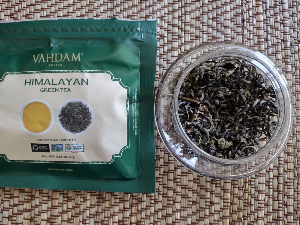 Close-up overhead shot of dry Himalayan green tea leaves in a clear glass jar, showcasing their dark green colour and somewhat twisted, large leaf structure. The Vahdam tea packet lies above the jar on a textured placemat.
