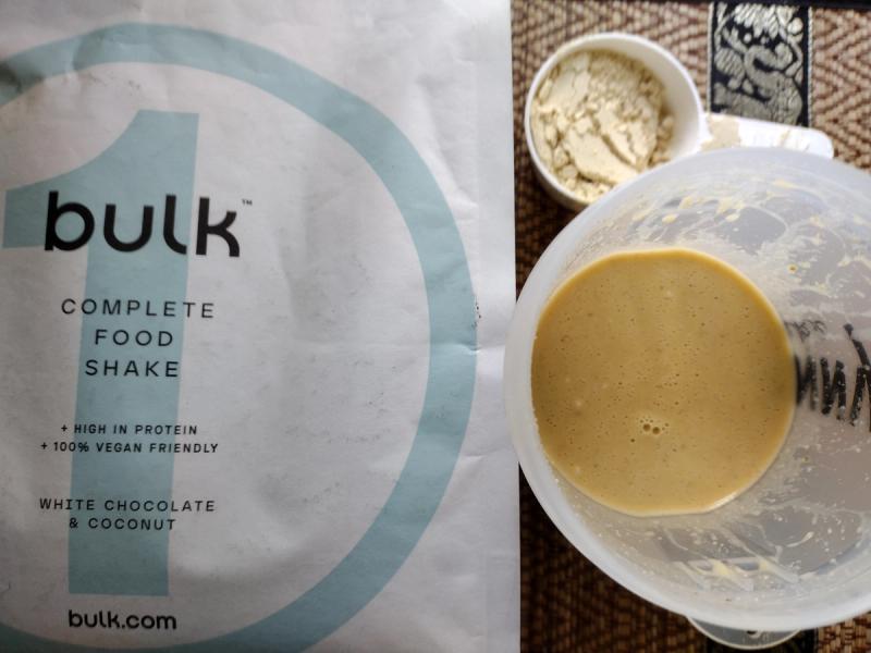 A prepared Bulk 1 Complete Food Shake showing its brownish-tan colour in a shaker, with the product pouch and a scoop of the raw powder alongside.