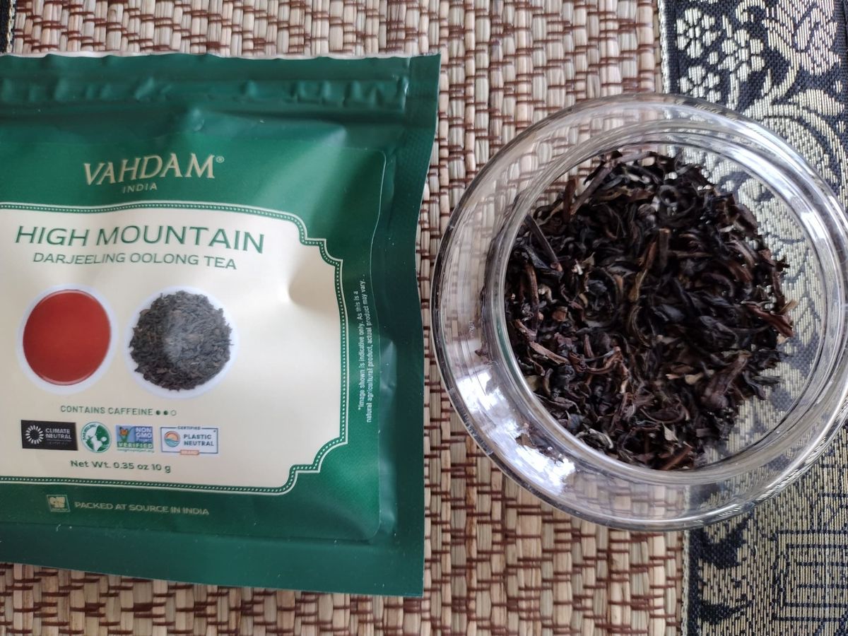 Overhead close-up of dry Darjeeling Oolong tea leaves in a clear jar. The leaves are dark brown/black, loosely twisted, and wiry. The green Vahdam tea packet lies next to the jar on a patterned surface.