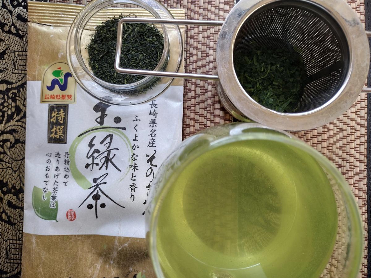 A Japanese green tea tasting setup featuring Saikaien Tokusen Tamaryokucha from Nagasaki Prefecture. The image shows a glass of cloudy, deep-steamed (fukamushi) green tea, vibrant emerald wet leaves in a stainless steel strainer, dark curled dry leaves in a glass dish, and the traditional Japanese packaging resting on a woven bamboo mat.