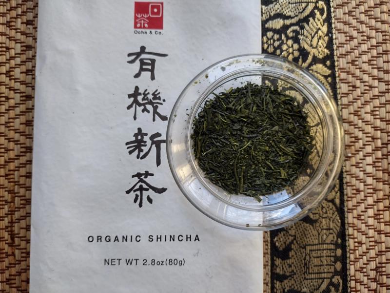 Ocha & Co Organic Shincha loose leaf green tea in a clear glass bowl displayed next to the white 80g packaging on a textured mat.