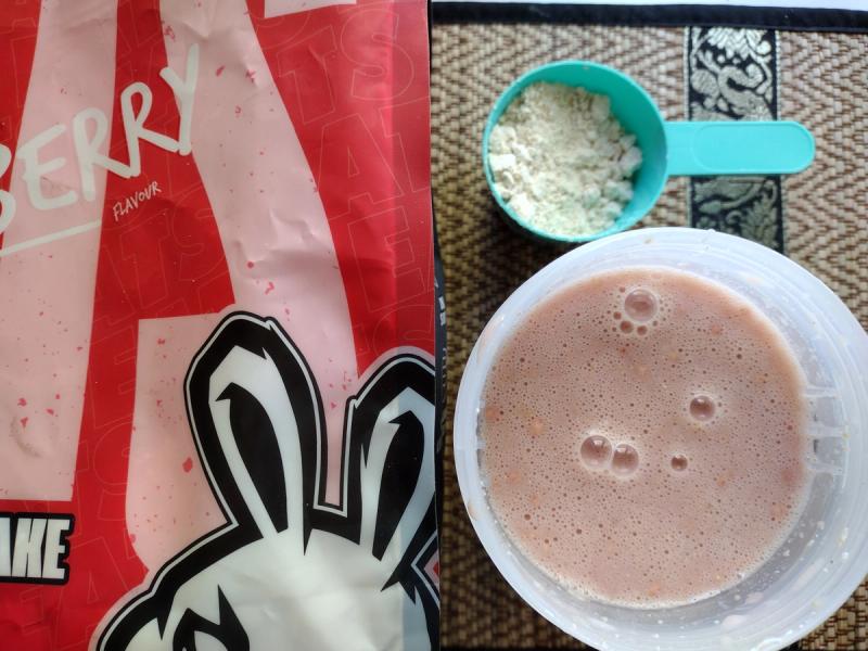 A scoop of Sneak Eats strawberry meal replacement powder next to the prepared pink shake in a shaker bottle, showing the texture and consistency for a product review.