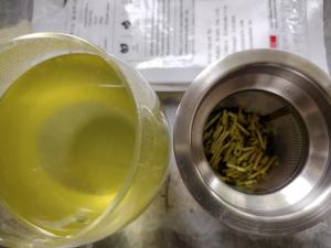A close-up shot of brewed Ocha & Co. Organic Kukicha. On the left, a glass contains the clear, bright, yellow-green tea liquor. On the right, a fine mesh metal strainer holds the infused, wet tea stems and leaves. The out-of-focus tea package is visible in the background.