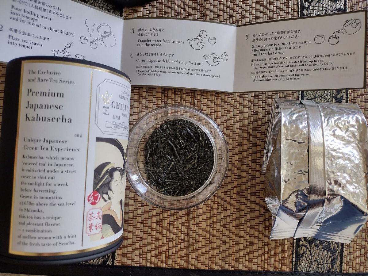 Dry leaves of Chill Tea Tokyo Kabusecha Hikari premium Japanese green tea in a glass jar, placed next to the black branded tin, silver inner foil pouch, and open brewing instruction leaflet on a table.