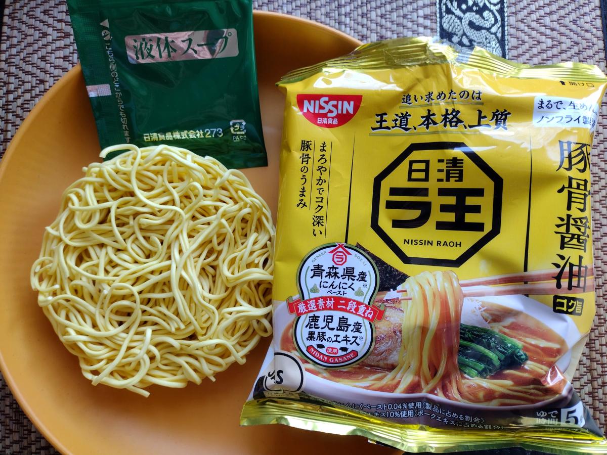 Uncooked contents of a Nissin Raoh Tonkotsu Shoyu ramen pack on an orange plate, displaying the yellow packaging, the air-dried non-fried noodle block, and the green liquid soup sachet.