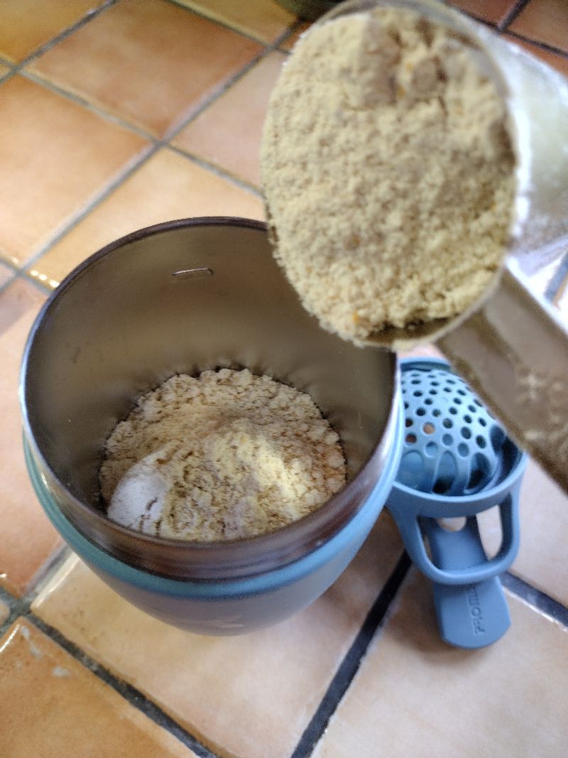 Preparing a shake by adding powder to a Promixx Pursuit insulated shaker, showing its stainless steel interior and blue snap-fit agitator on a tiled counter.