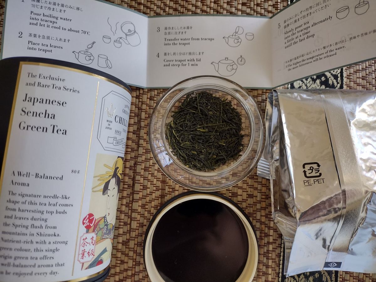 Dry loose leaves of Chill Tea Tokyo Sencha Midori in a clear jar. The leaves are dark green and needle-shaped. Next to the jar is the open black tea canister, the silver foil inner pouch, and brewing instructions