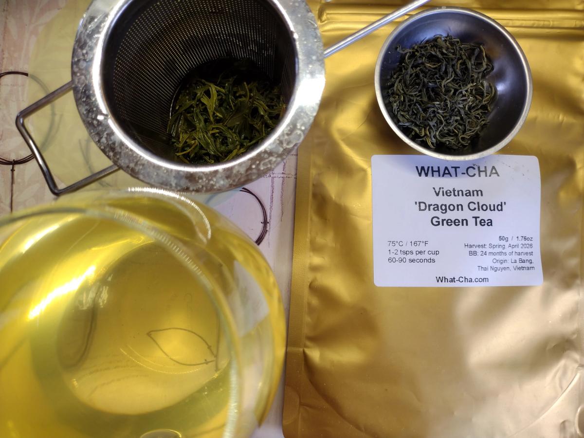 A glass cup of brewed What-Cha Vietnam Dragon Cloud Green tea with a golden-yellow liquor, next to a metal strainer of bright green wet leaves, a bowl of dark wiry dry leaves, and the original label on gold packaging.