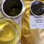 A glass cup of brewed What-Cha Vietnam Dragon Cloud Green tea with a golden-yellow liquor, next to a metal strainer of bright green wet leaves, a bowl of dark wiry dry leaves, and the original label on gold packaging.