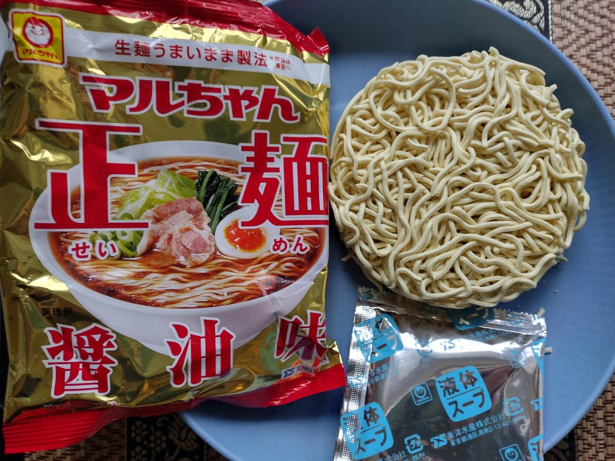 A gold packet of Maruchan Seimen Shoyu instant ramen next to a perfectly round nest of air-dried noodles and a silver liquid soup sachet on a blue plate.