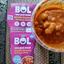Open pot of BOL Middle Eastern Harissa Stew showing a spoon lifting white beans and chickpeas in a thick tomato sauce, with pink packaging in the background.