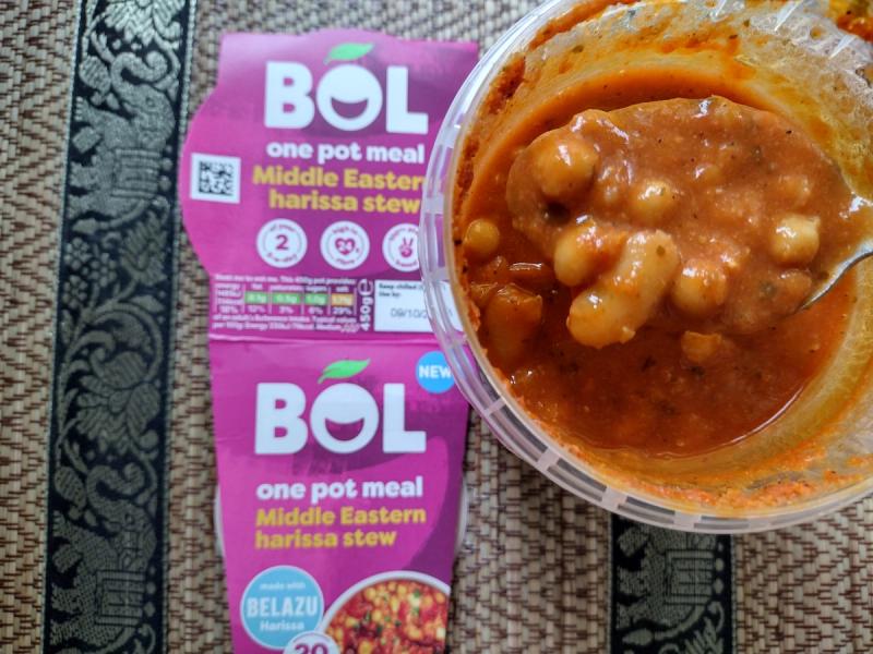 Open pot of BOL Middle Eastern Harissa Stew showing a spoon lifting white beans and chickpeas in a thick tomato sauce, with pink packaging in the background.