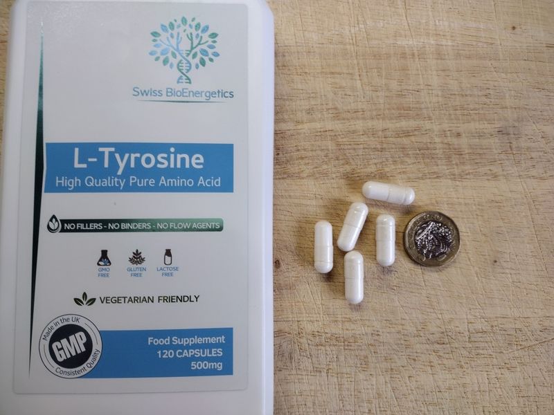 A white bottle of Swiss BioEnergetics L-Tyrosine on a wood surface, next to five white 500mg capsules and a £1 coin to show their size