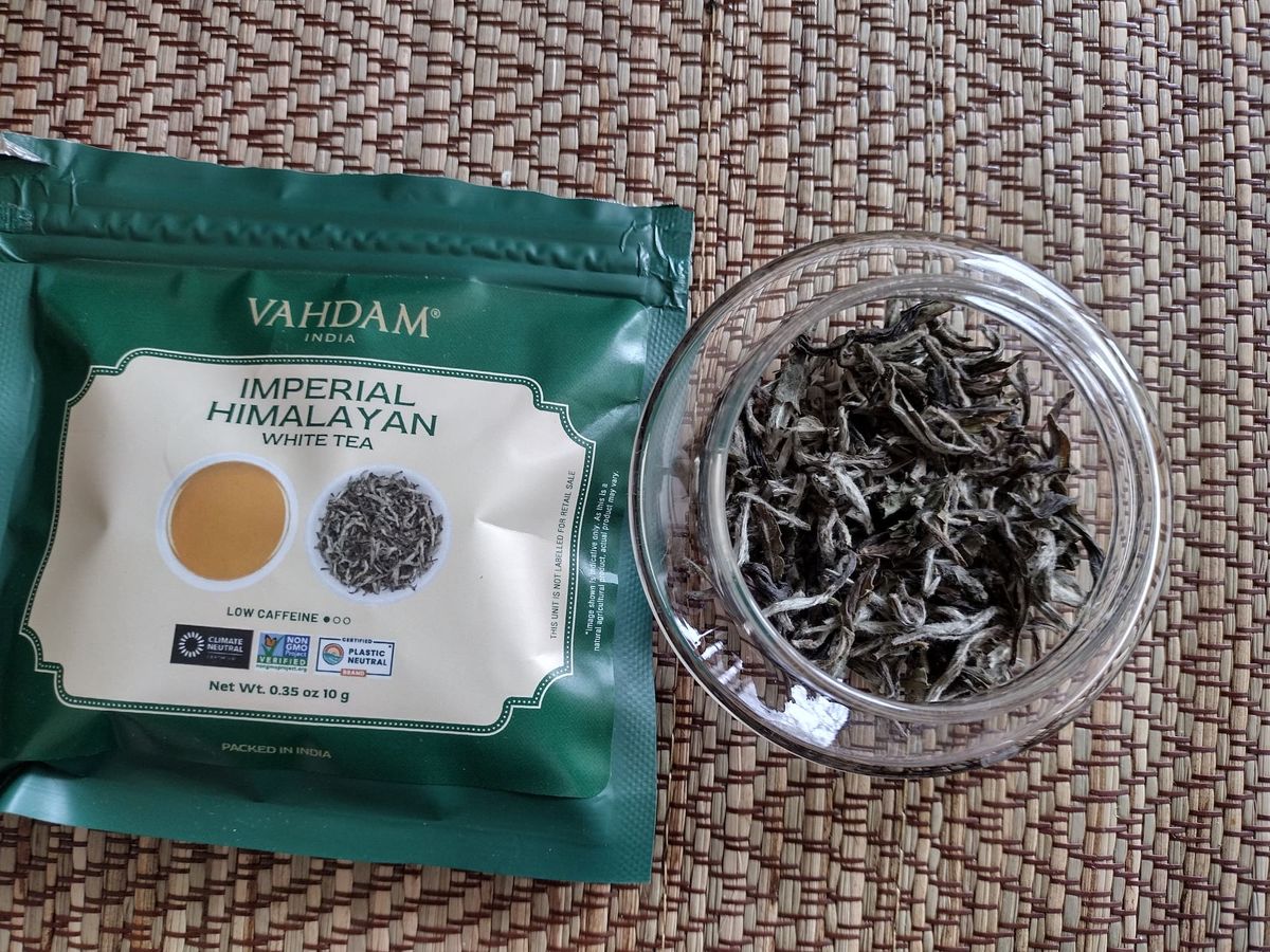 Overhead close-up of dry Imperial Himalayan white tea leaves in a small clear jar. The leaves are greenish-grey with visible fuzzy silver buds, appearing loose and delicate. The green Vahdam tea packet lies beside the jar on a textured placemat.
