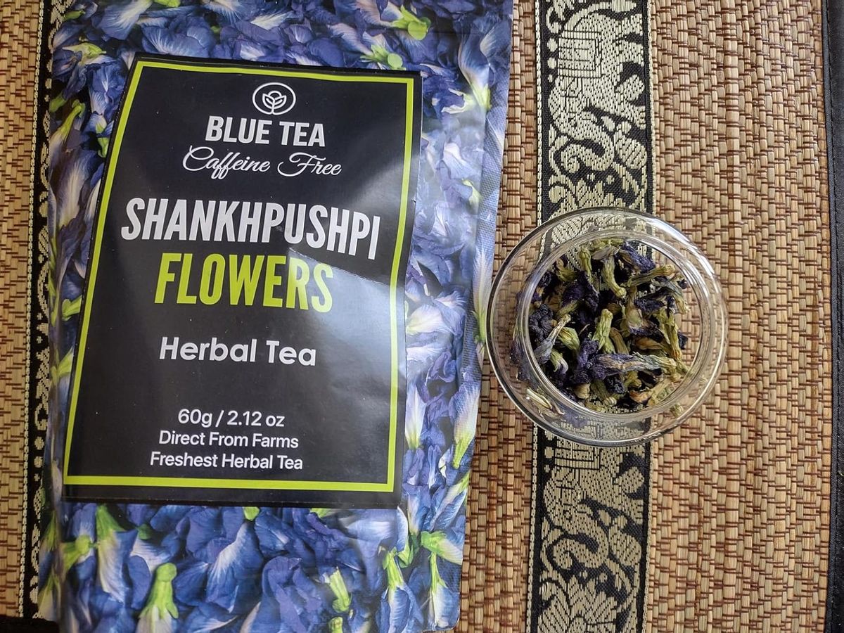 This clearly shows the "BLUE TEA Shankhpushpi Flowers" packaging (60g pouch, mentions "Direct From Farms", "Freshest Herbal Tea", "Caffeine Free"). It also shows the dried flowers themselves in a separate jar, giving a good look at their appearance and condition (they look like whole, well-dried flowers).