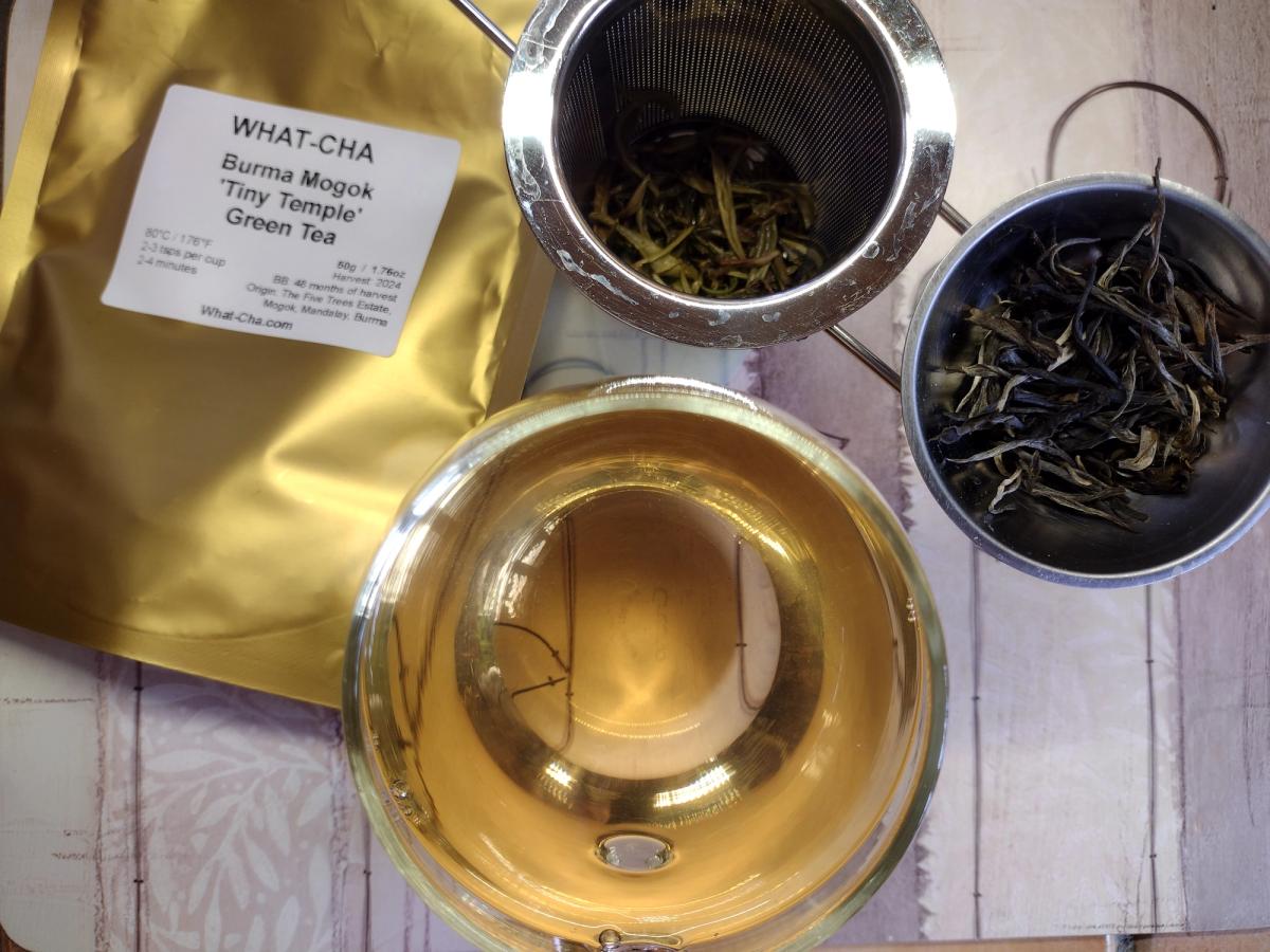 What-Cha Burma Mogok 'Tiny Temple' loose leaf green tea showing the gold packaging, wiry dry leaves, steeped wet leaves in a metal strainer, and a glass cup of clear, golden-yellow brewed tea.