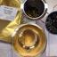 What-Cha Burma Mogok 'Tiny Temple' loose leaf green tea showing the gold packaging, wiry dry leaves, steeped wet leaves in a metal strainer, and a glass cup of clear, golden-yellow brewed tea.