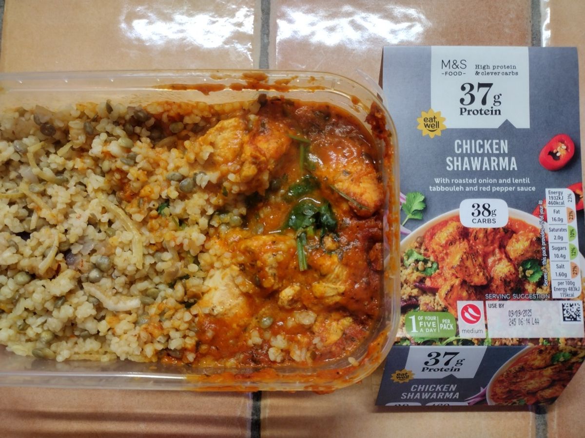 An overhead review photo of the M&S High Protein Chicken Shawarma. The prepared meal, with chicken in a red pepper sauce and a side of lentil tabbouleh, is shown in its container next to the product packaging.
