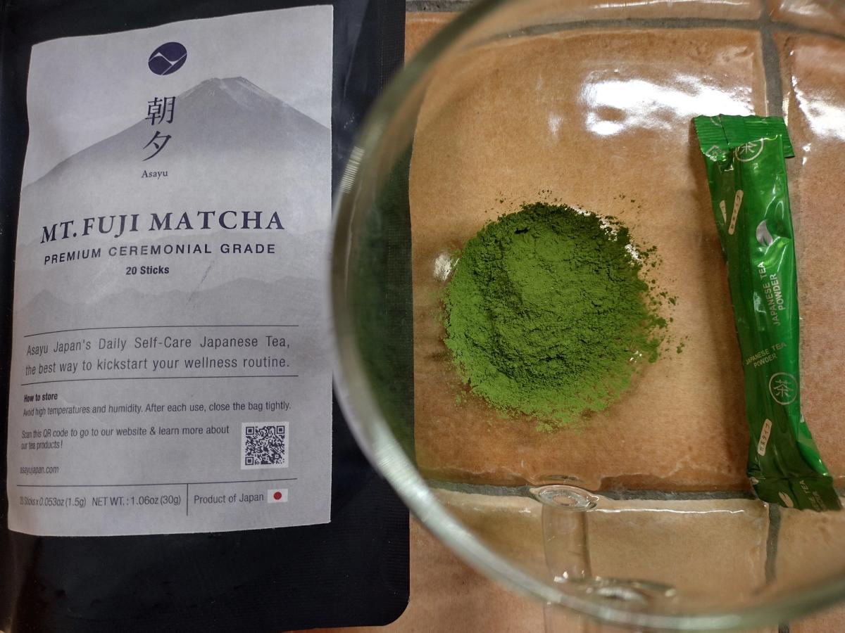Asayu Japan Mt. Fuji Matcha packaging next to a glass bowl containing a mound of vibrant green, clumpy ceremonial grade powder and an individual single-serve stick.