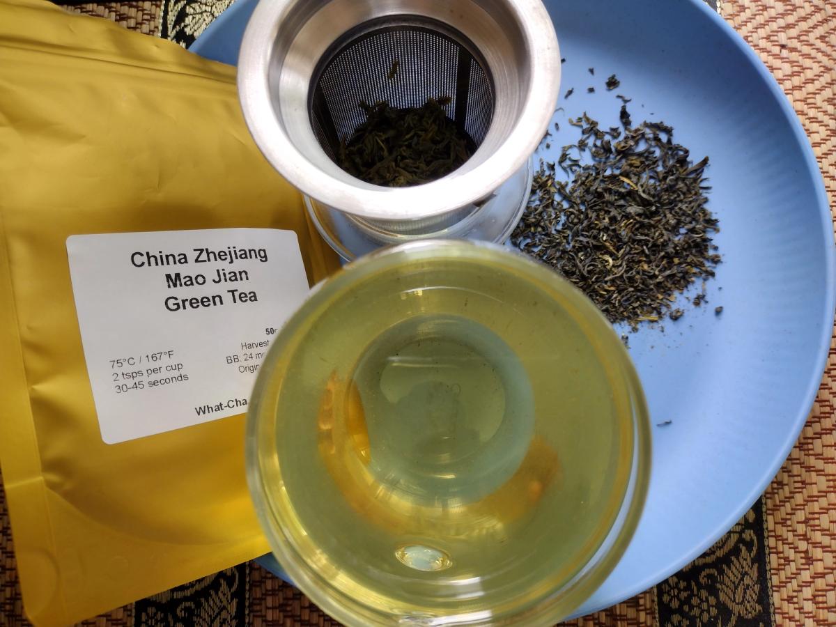 A freshly brewed glass cup of What-Cha China Zhejiang Mao Jian loose leaf green tea, displayed on a blue plate alongside dry tea leaves, a stainless steel infuser with wet leaves, and the yellow packaging bag.