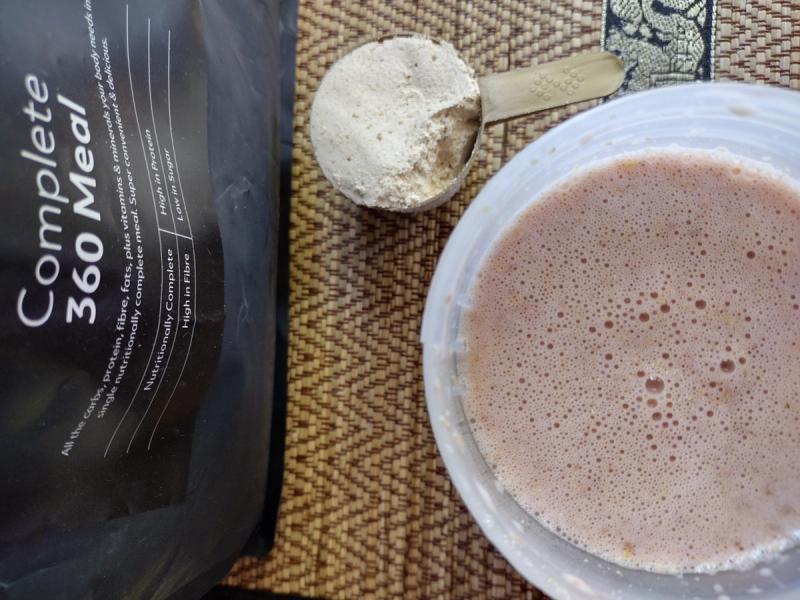 A shaker cup with The Protein Works Complete 360 Meal mixed with water, next to the product pouch and a scoop of the Strawberries & Cream powder.