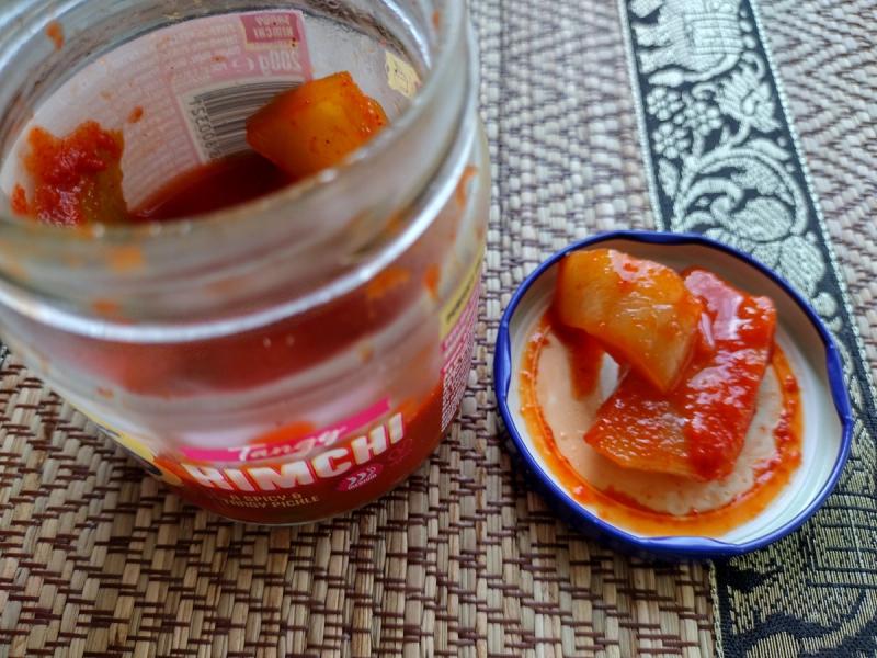 Open jar of Blue Dragon Tangy Kimchi Topper on a woven mat, showing the blue lid with large chunks of mustard leaf coated in a thick, glossy red chilli sauce.