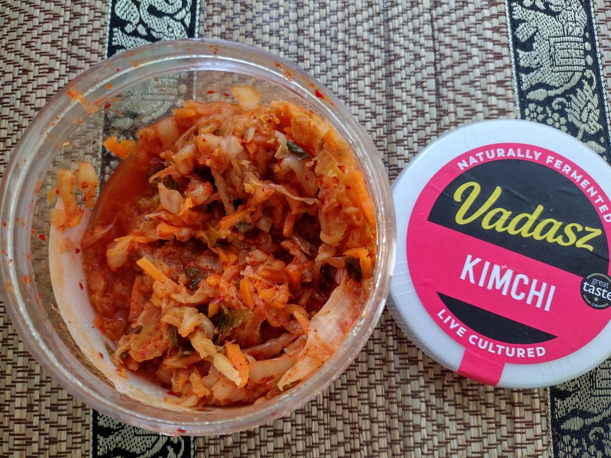 An open tub of Vadasz Kimchi, a popular live cultured and naturally fermented food in the UK. The fresh vegan kimchi is shown next to the pink Vadasz branding on the lid.