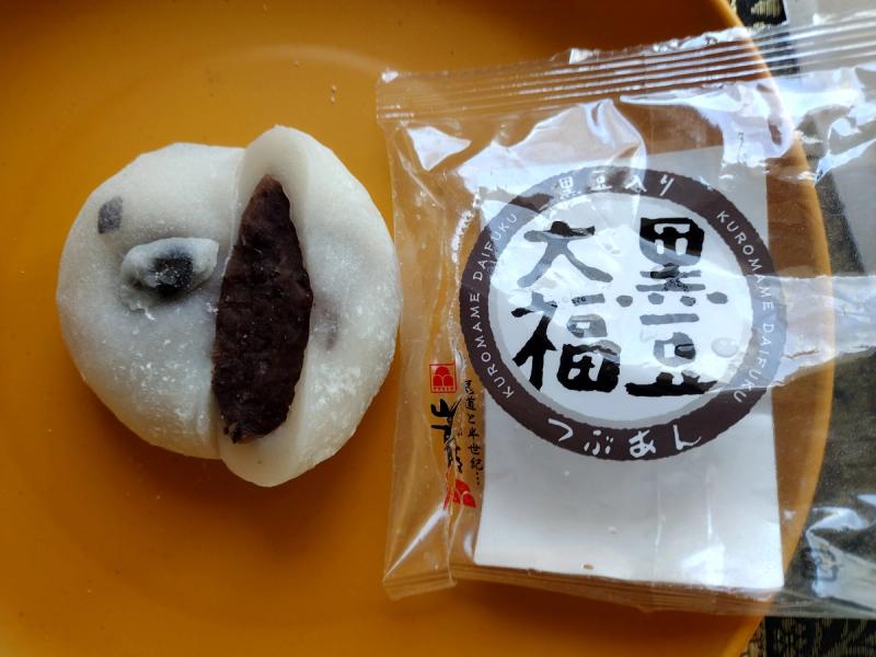 Cross section of a Yamamotoya Kuromame Daifuku on a yellow plate showing the thick white mochi skin embedded with whole black soybeans and a chunky red bean paste centre, alongside the open Japanese packaging.