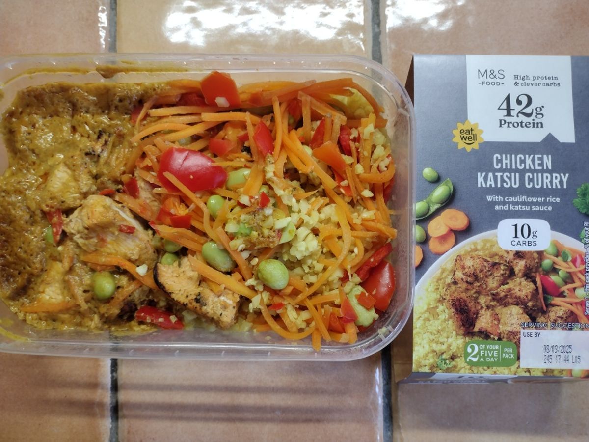 Overhead review photo of the M&S High Protein Chicken Katsu Curry. The prepared low-carb meal with cauliflower rice and chicken is shown in its container next to the product's packaging.