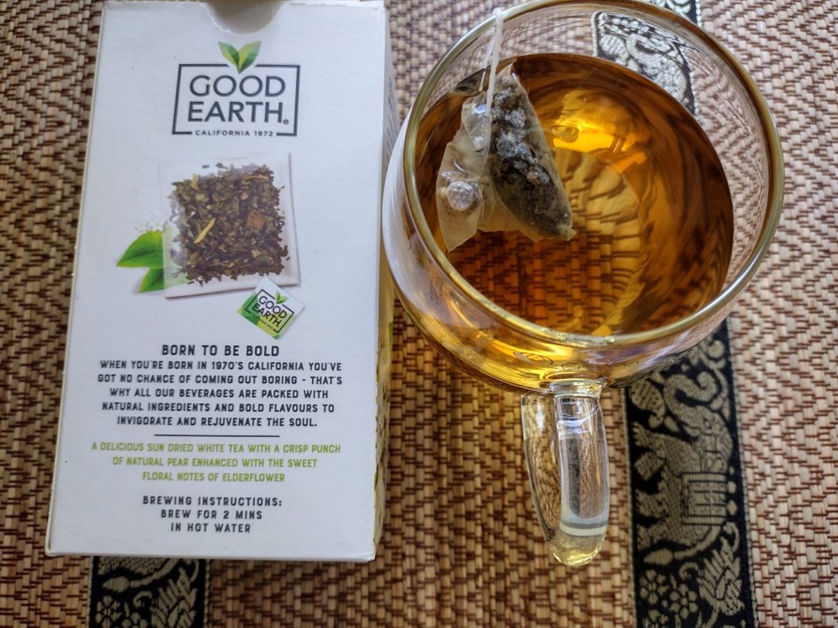 Good Earth Born to be Bold white tea brewed in a clear glass cup, showing its amber colour. The pyramid teabag steeps inside, alongside the product box describing the pear and elderflower flavour.