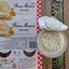 Overhead view of a Bonne Maman Clementine and Lemon Mousse cardboard sleeve next to an opened plastic pot revealing a highly aerated, pale orange citrus yoghurt mousse on a woven table mat.
