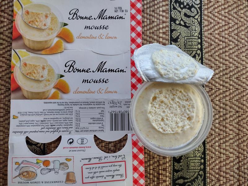 Overhead view of a Bonne Maman Clementine and Lemon Mousse cardboard sleeve next to an opened plastic pot revealing a highly aerated, pale orange citrus yoghurt mousse on a woven table mat.