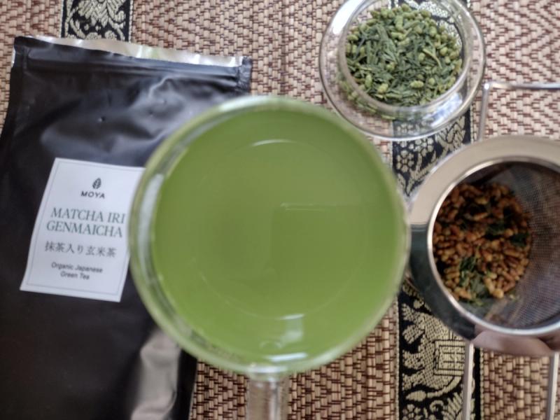 A vibrant green cup of brewed Moya Matcha Iri Genmaicha, with the dry tea leaves and roasted brown rice shown in a dish and strainer.