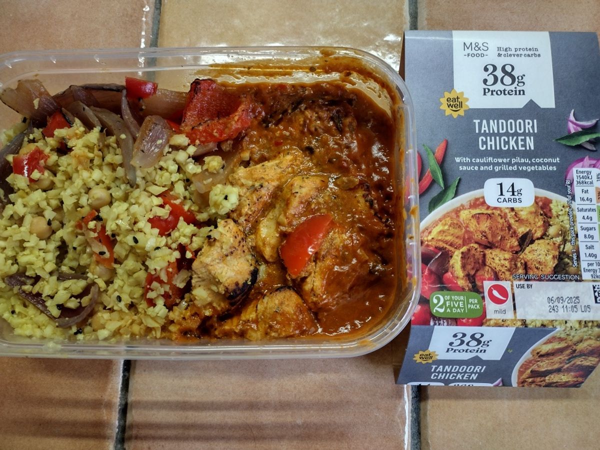 An overhead review photo of the M&S High Protein Tandoori Chicken. The prepared low-carb meal, showing the chicken in sauce and the cauliflower pilau, is in its tray next to the product packaging.