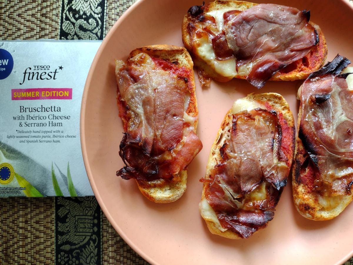 A close-up shot of four golden-brown bruschetta topped with tomato purée, melted cheese, and crisped Serrano ham on a stoneware plate, with the corner of the Tesco Finest packaging visible.