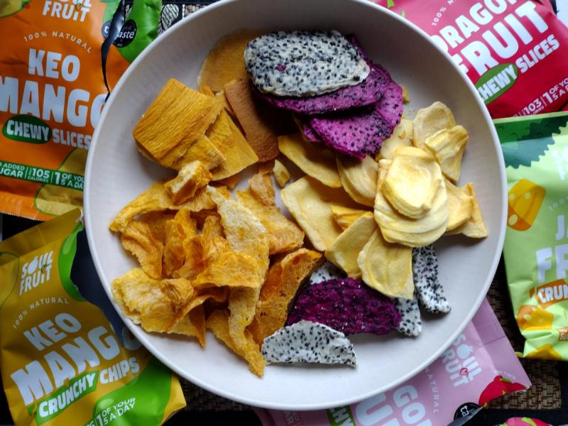 A white bowl filled with an assortment of Soul Fruit brand dried snacks, featuring crunchy purple and white freeze-dried dragon fruit chips, flat soft-dried mango slices, irregular freeze-dried mango chunks, and yellow jackfruit chips. Colorful Soul Fruit packaging bags for Keo Mango and Dragon Fruit surround the bowl.