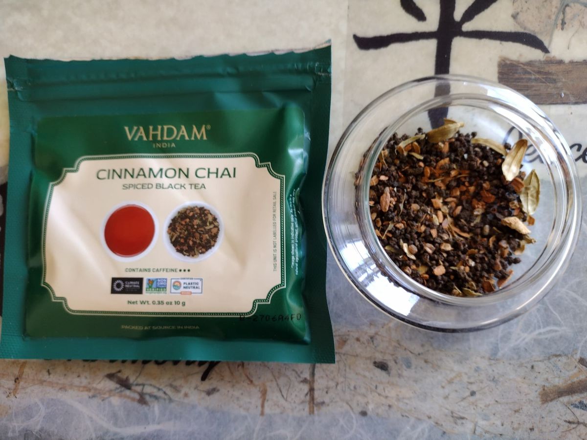 Overhead close-up of the dry Cinnamon Chai mix in a clear jar. Dark brown CTC tea granules are mixed with visible whole green cardamom pods, pieces of cinnamon bark, and other lighter spice fragments. The green Vahdam tea packet is beside it.