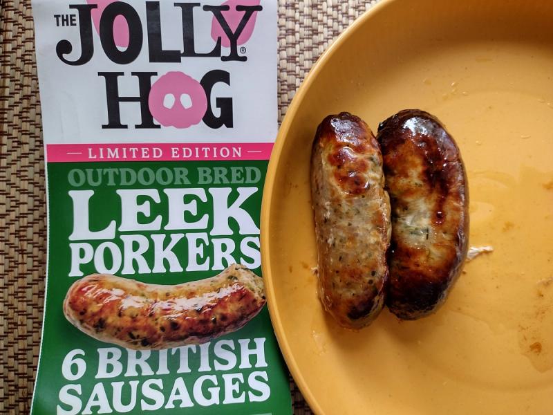 A review photo of The Jolly Hog's Limited Edition Leek Porker sausages, showing two cooked and greasy sausages on a yellow plate next to the green product packaging.