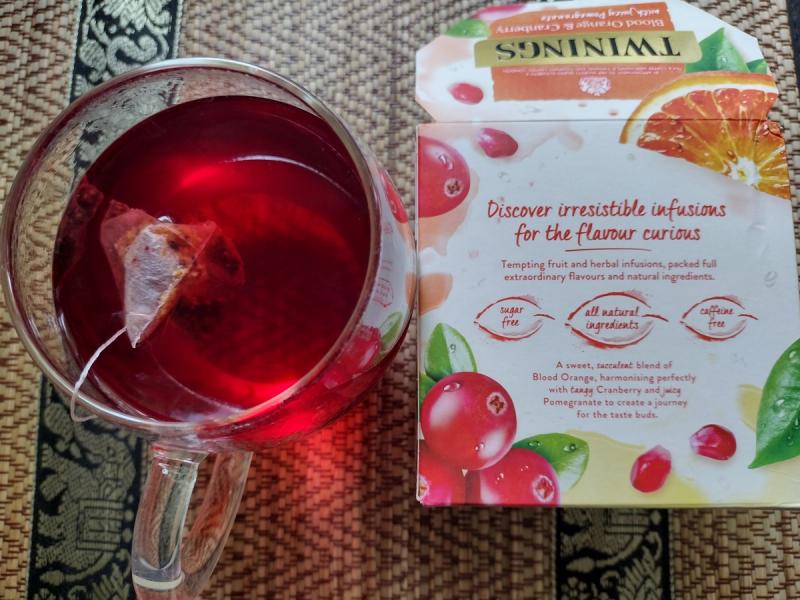 A glass mug of vibrant red Twinings Blood Orange & Cranberry fruit infusion steeping next to its product box.