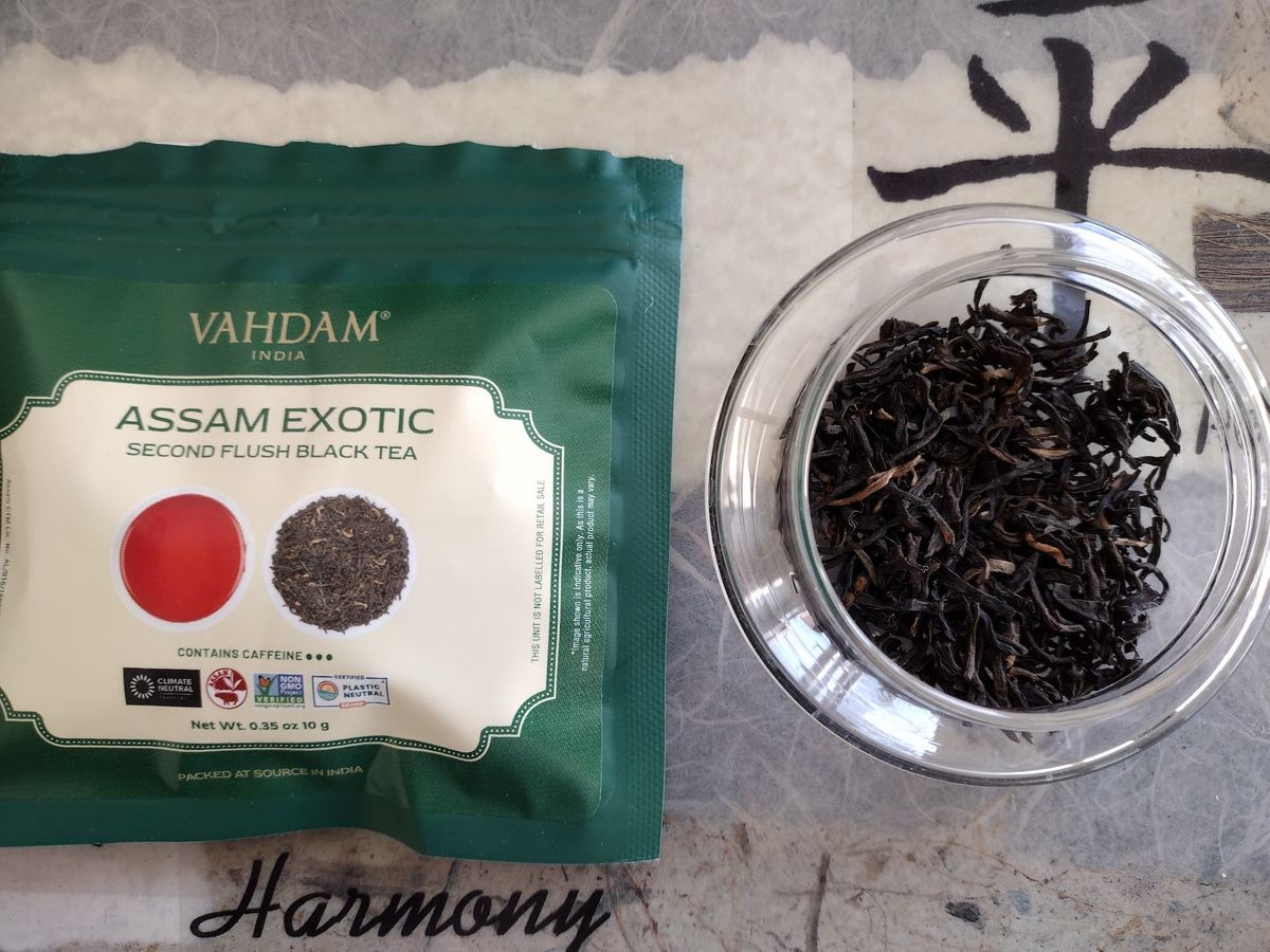 Overhead close-up of dry Assam Exotic black tea leaves in a clear jar. The leaves are dark brown to black, wiry, with noticeable interspersed golden tips (buds). The green Vahdam tea packet lies beside the jar on a textured surface.