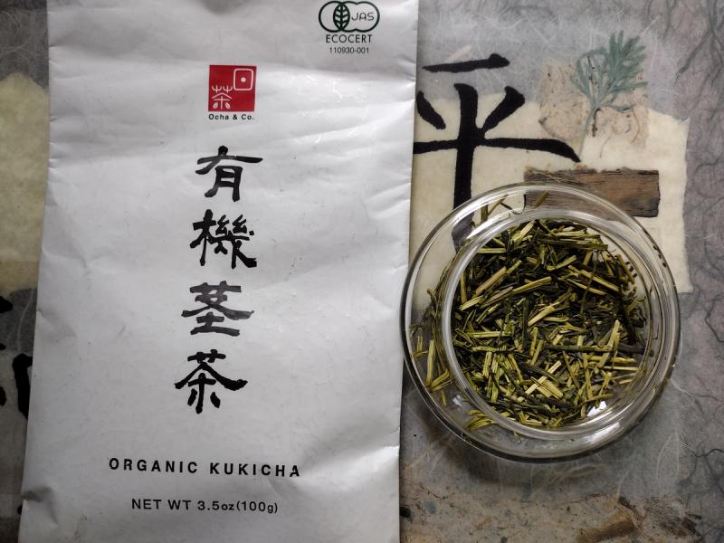 A flat lay photo of Ocha & Co. Organic Kukicha Japanese twig tea. On the left is the white vacuum-sealed package with Japanese calligraphy for 'Yuki Kukicha' and the JAS and ECOCERT organic certification logos. On the right, a small glass bowl displays the dry tea, showing a mix of light green stems and darker green leaf fragments.