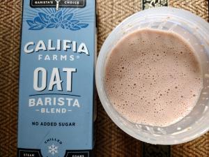 A prepared shake of The Protein Works Complete 360 Meal in Strawberries & Cream flavour next to the carton of Califia Farms Oat Barista Blend.