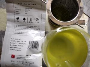 Detailed view of the back label of Ocha & Co. Fukamushicha, listing Japan as the origin and brewing parameters (75-85°C for 30-40 sec). Next to it is a glass of the prepared vibrant, opaque green tea, with the tea strainer resting on top.