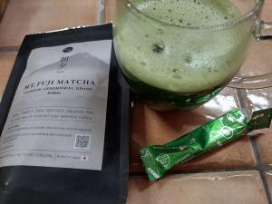 Brewed Asayu Mt. Fuji ceremonial matcha in a glass cup showing airy bubbles and froth, placed next to the empty green stick wrapper and black product pouch.