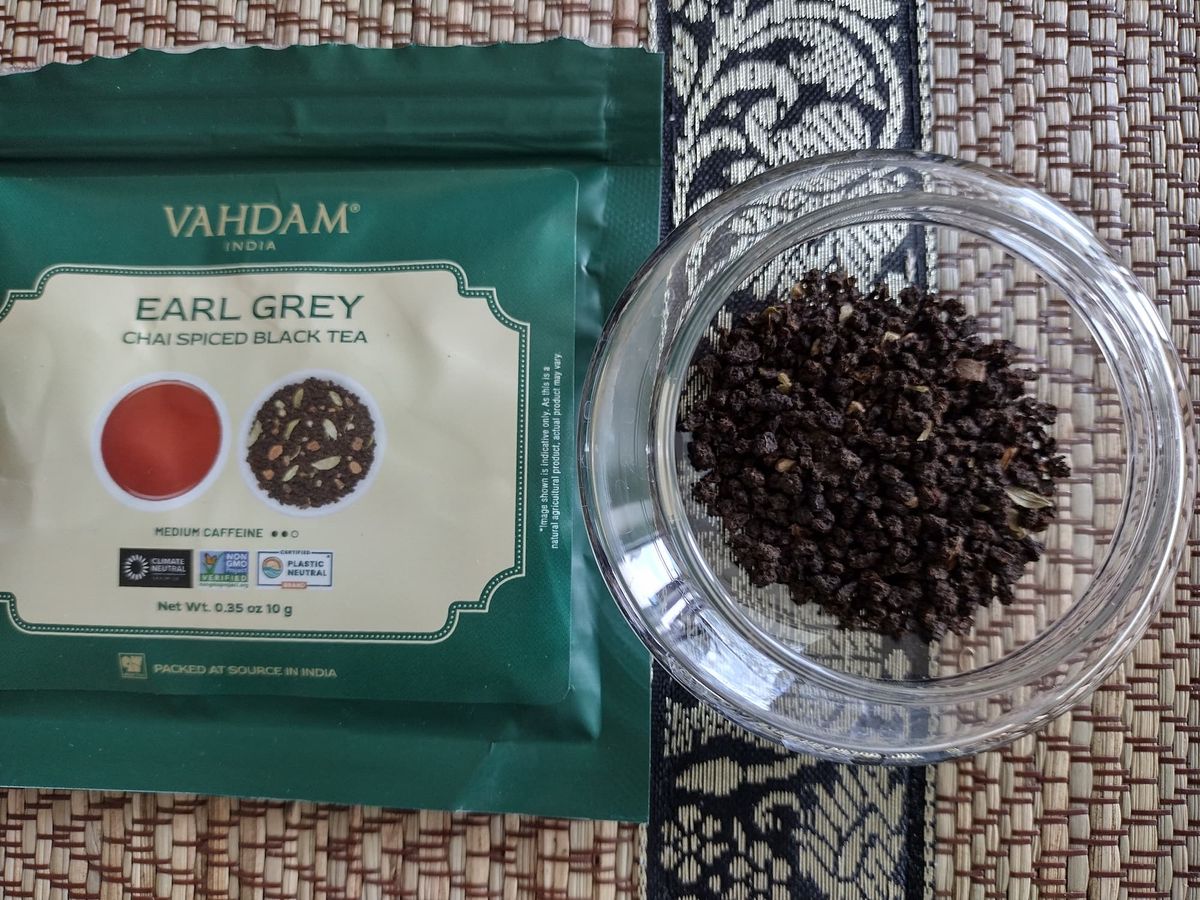 Overhead close-up of the dry Earl Grey Chai Spiced Black Tea in a clear jar. The blend primarily consists of small, dark brown CTC tea granules mixed with some visible lighter-coloured spice fragments. The green Vahdam tea packet is positioned next to the jar on a patterned placemat.