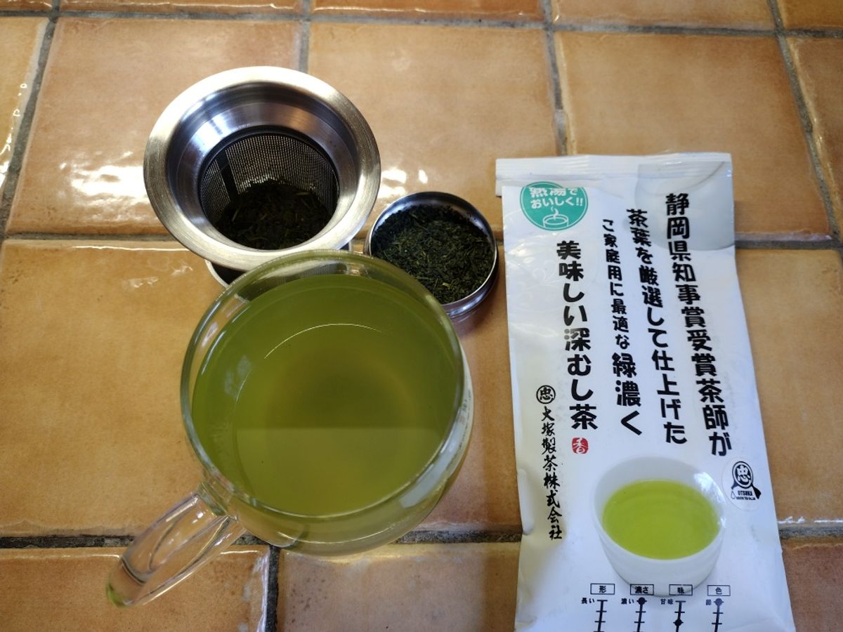 A flat lay of Otsuka Fukamushi tea showing the fine, broken dry leaves, the vibrant cloudy green liquor in a mug, and the steeped leaves in a metal infuser, alongside the original Japanese packaging.