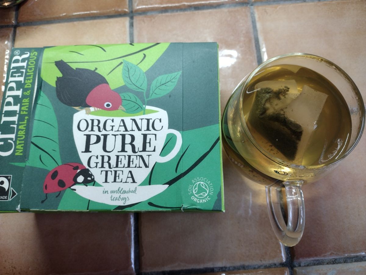 A box of Clipper Organic Pure Green Tea with a glass mug showing the light, yellow-green colour of the brewed tea.