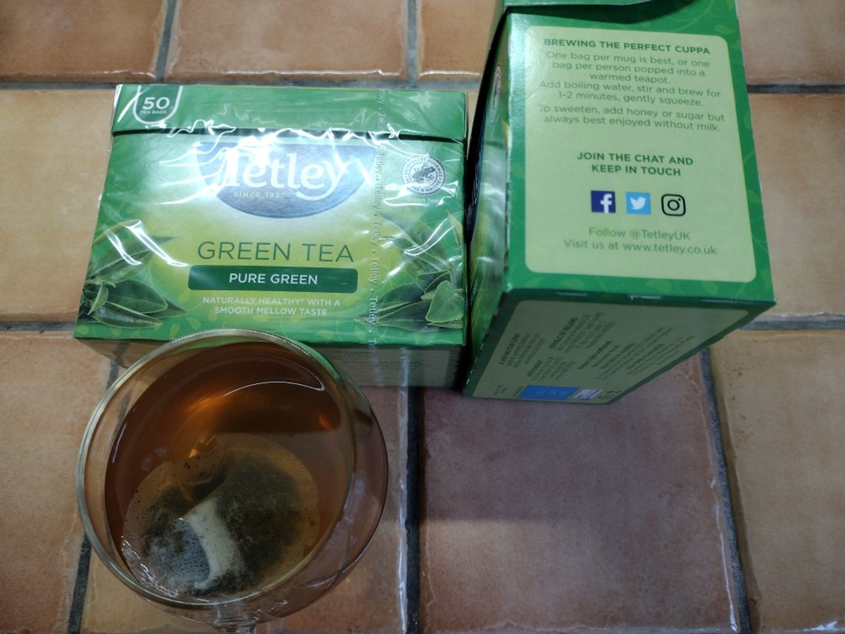 A green box of Tetley Pure Green Tea next to a glass mug containing the dark, yellow-brown brewed tea.